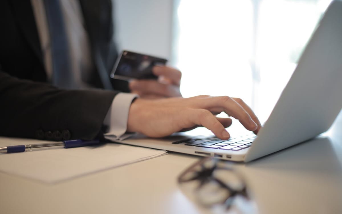A seller at a laptop holding a payment card mid-checkout, glasses and pen on the desk — managing cross-border payments and supplier settlements for a dropshipping business