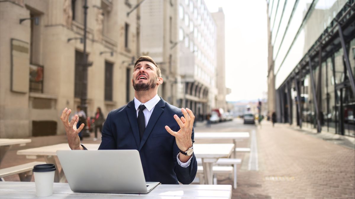 A businessman in a suit at an outdoor café table with his laptop, looking up in exasperation — the moment a bank rejection lands