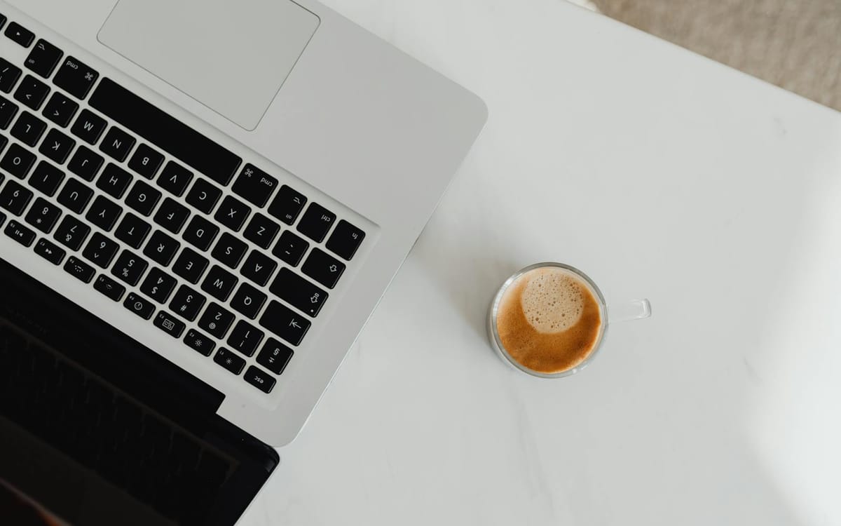 Top-down view of an open laptop beside a glass cup of coffee on a clean white desk — a borderless business run from a single device