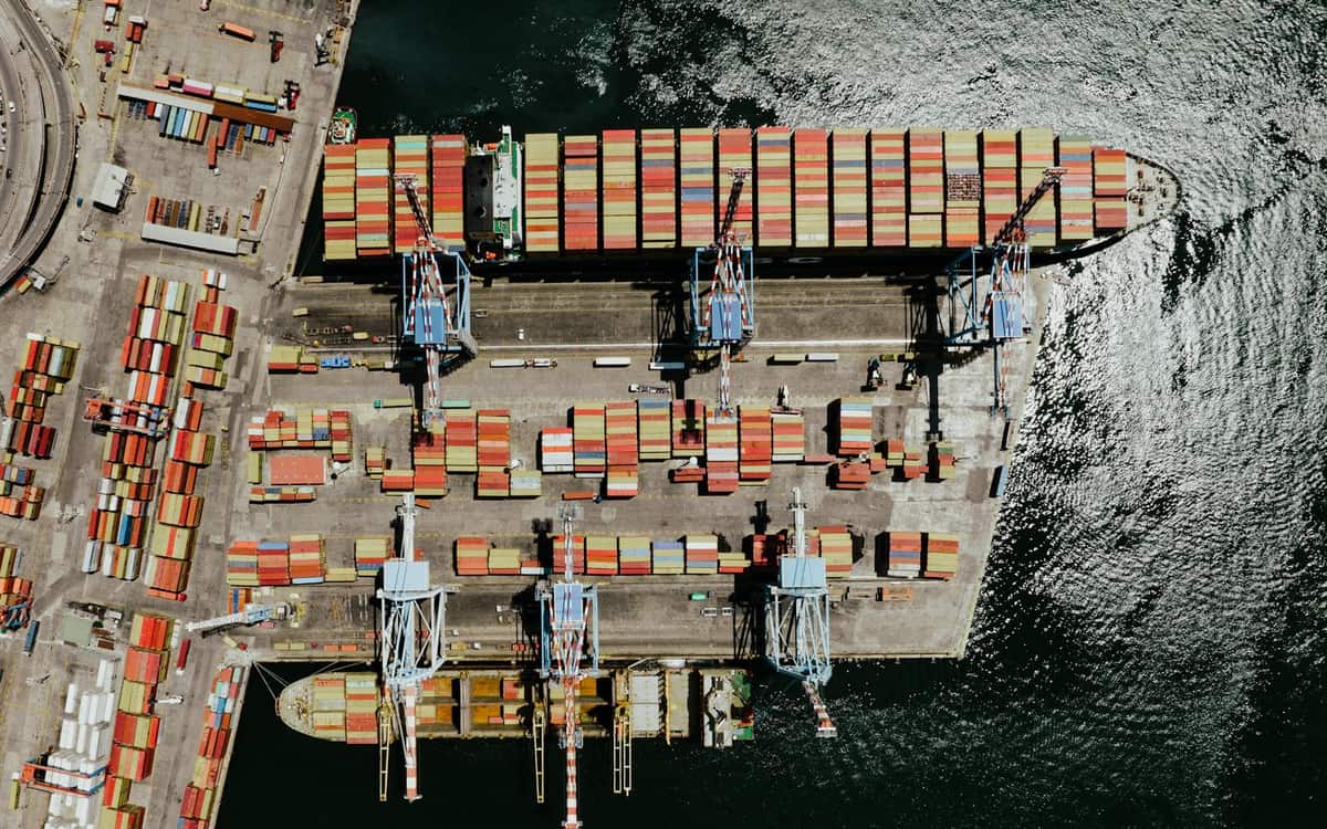 Aerial view of a large container ship berthed at a quay being loaded by gantry cranes, with stacked containers on the dockside — the freight flows a Hong Kong trading company settles and finances