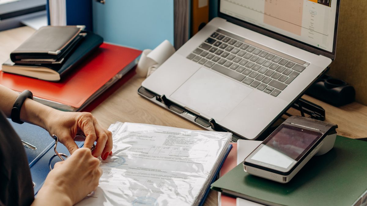 A desk with lever-arch files, a laptop and a card-payment terminal as a document is reviewed — the real operations and record-keeping of a genuine business, not a brass-plate shell