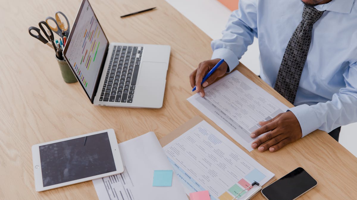 A founder at a desk with a laptop showing project charts, a tablet and printed reports — planning the operations of a remotely-run company
