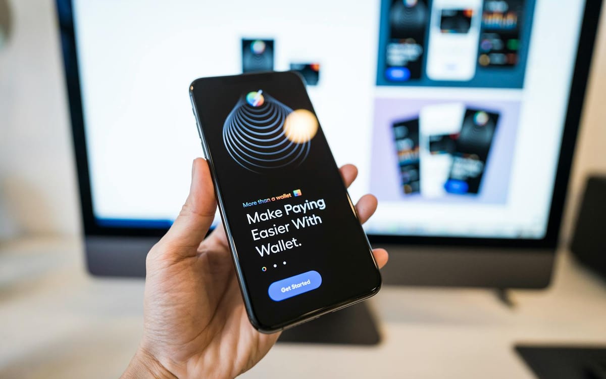 A hand holding a smartphone showing a digital payments wallet app, with app screens on a monitor behind — the multi-currency banking and payments layer a solo consultant runs on a Hong Kong company